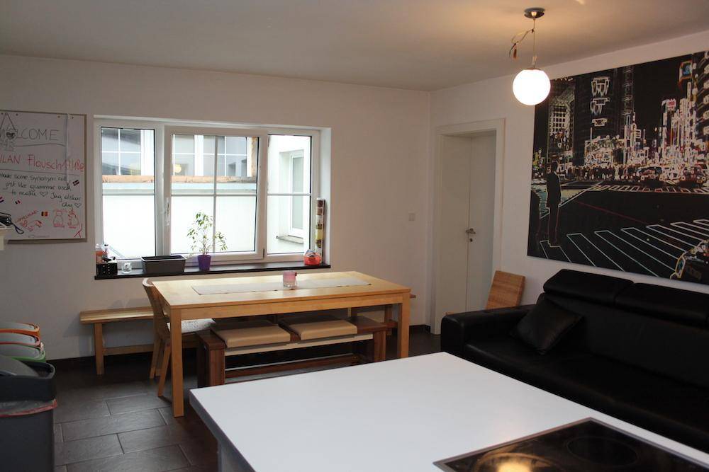 Offener Wohn- und Essbereich mit Holztisch, schwarzem Sofa und großem Fenster, ausgestattet mit dunklem Boden.