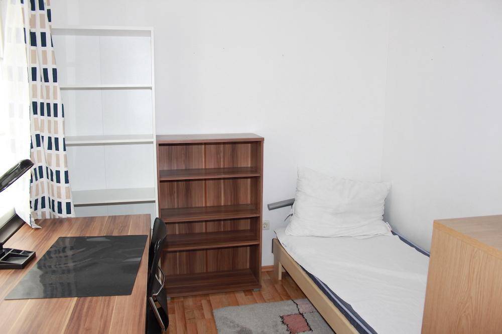 Einfaches Schlafzimmer mit Einzelbett, Schreibtisch und Bücherregalen, ausgestattet mit Parkettboden.