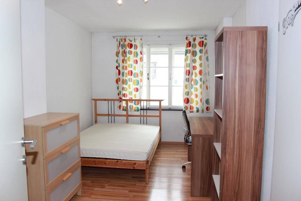 Helles Schlafzimmer mit Holzbett, Schreibtisch und bunten Vorhängen, ausgestattet mit Parkettboden.