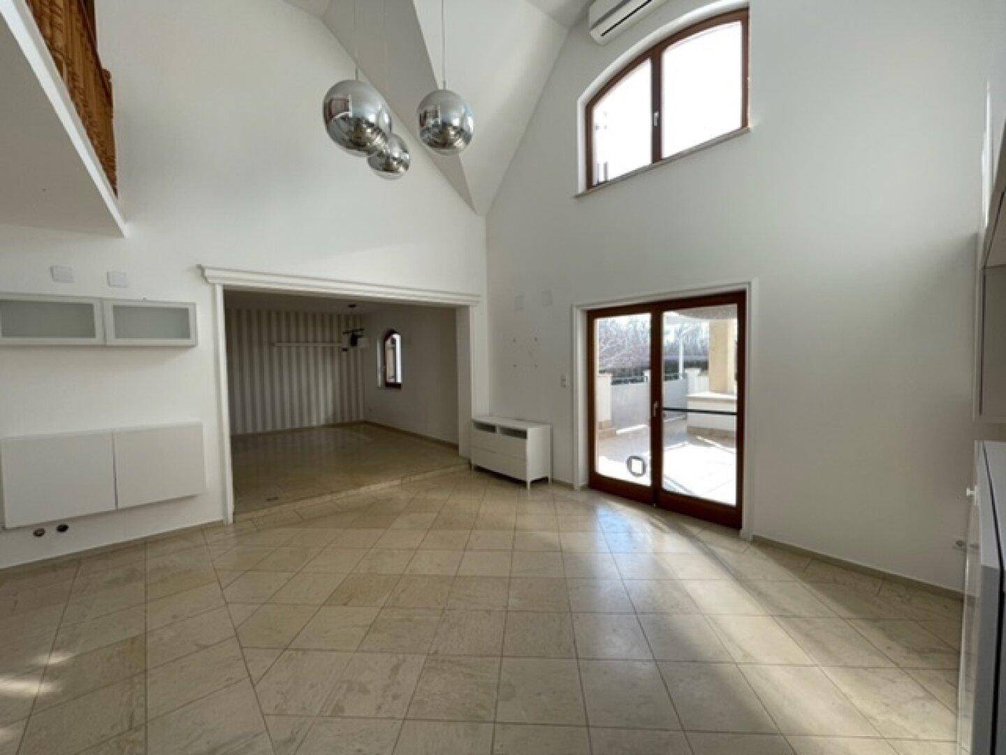 Heller Wohnbereich mit hohen Decken, Fliesenboden und Zugang zu einer Außenterrasse.