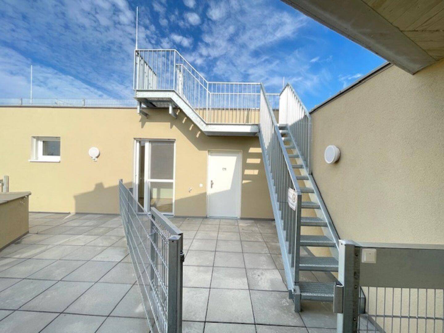 Dachterrasse mit Metalltreppe und Zugang zu weiteren Bereichen unter blauem Himmel.