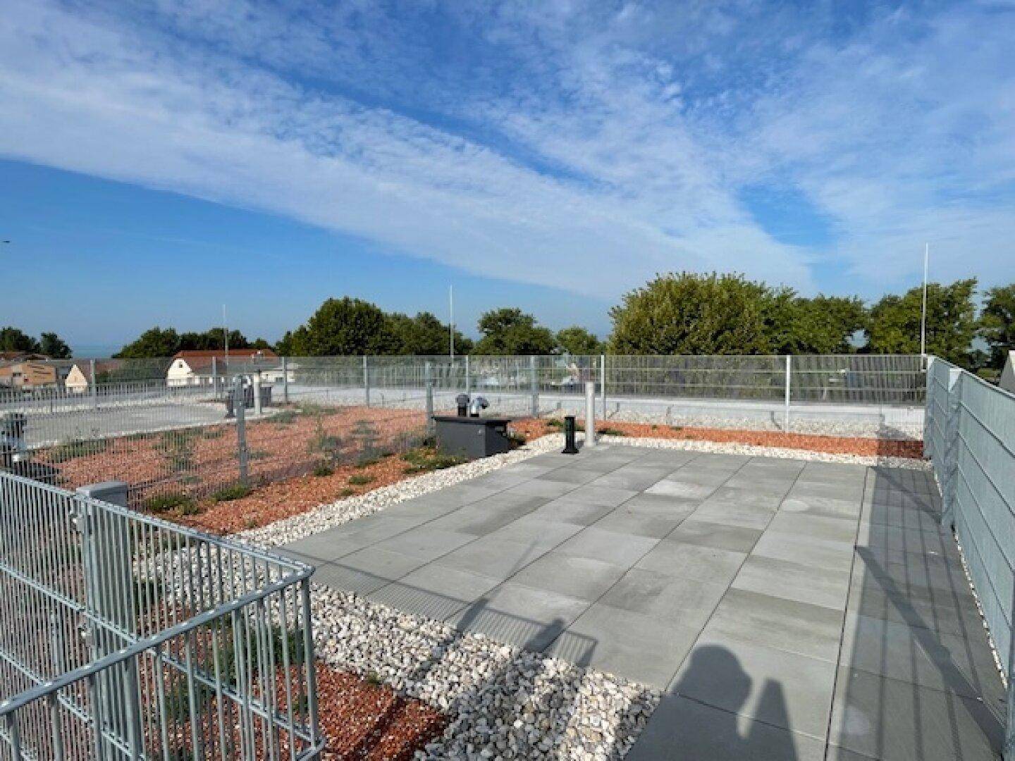 Geräumige Dachterrasse mit gepflasterten Flächen, Zierkies und niedriger Bepflanzung unter blauem Himmel.