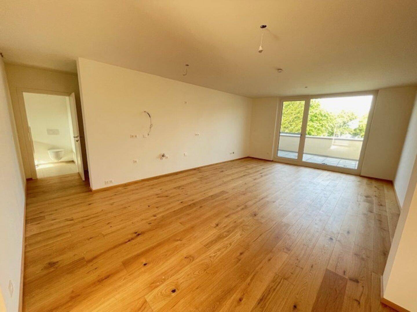 Geräumiges Wohnzimmer mit hellem Holzboden und Zugang zu einem Balkon, daneben ein Badezimmer.