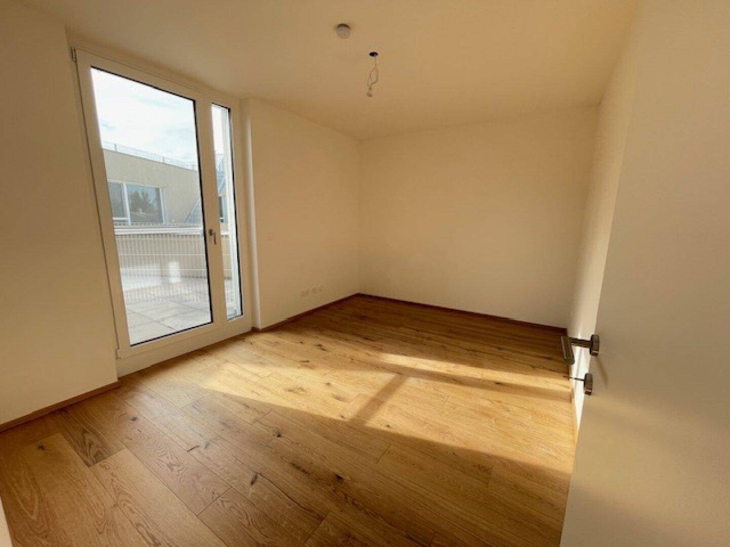 Helles Zimmer mit Holzboden und großer Glastür, die zu einem Außenbereich führt.