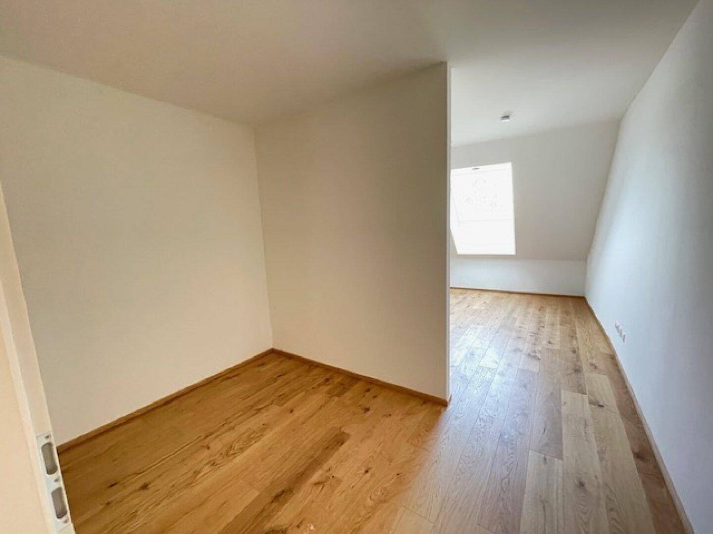 Helles Zimmer mit Holzboden und weiß gestrichenen Wänden, angrenzend an einen Raum mit Dachfenster.