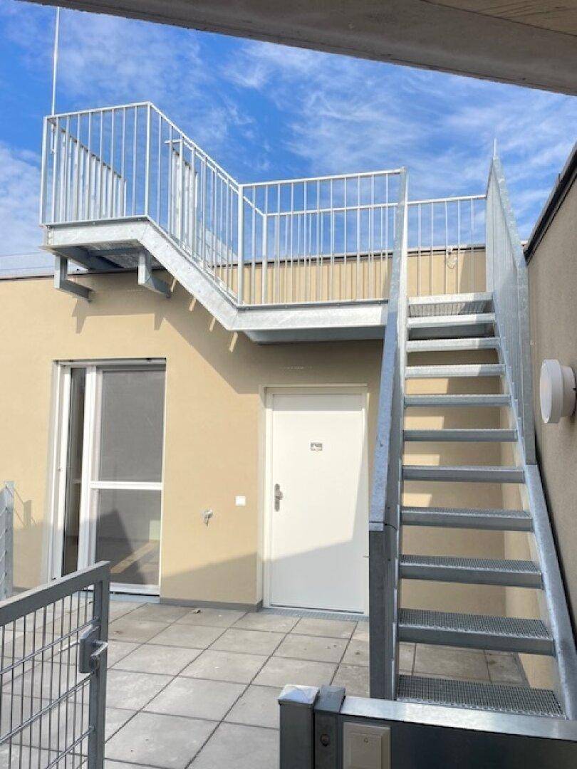 Dachterrasse mit Metalltreppe und Zugangstür, umgeben von einer schützenden Brüstung.