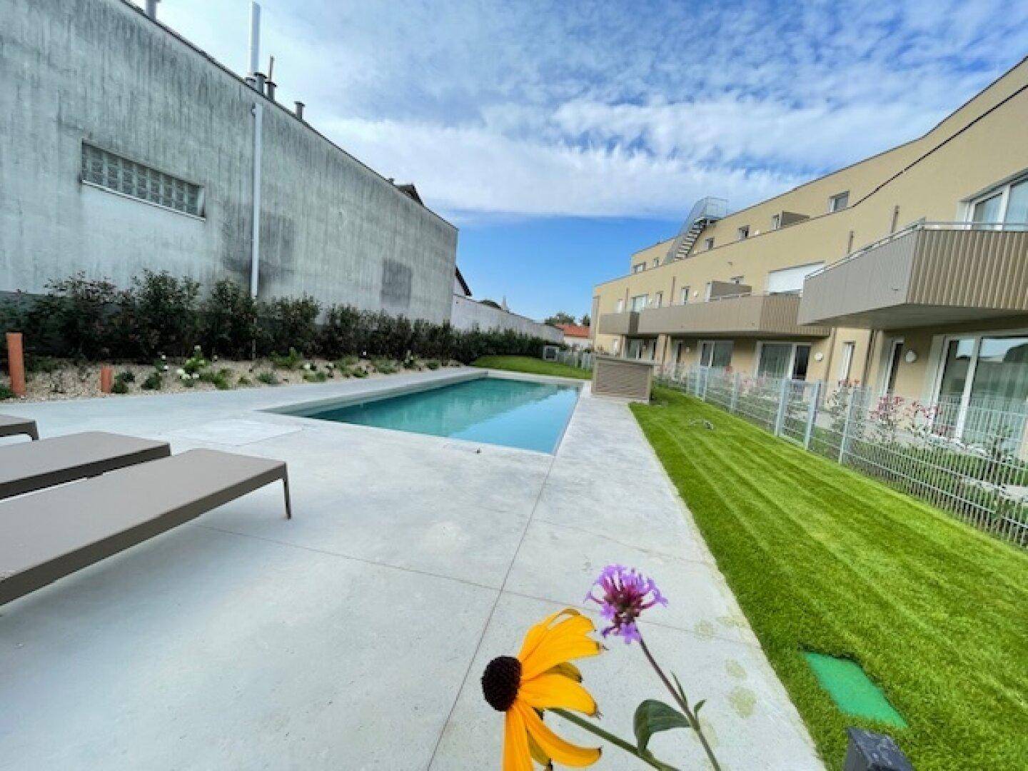Außenpoolbereich mit Liegestühlen, Rasenfläche und Blick auf die moderne Wohnanlage.