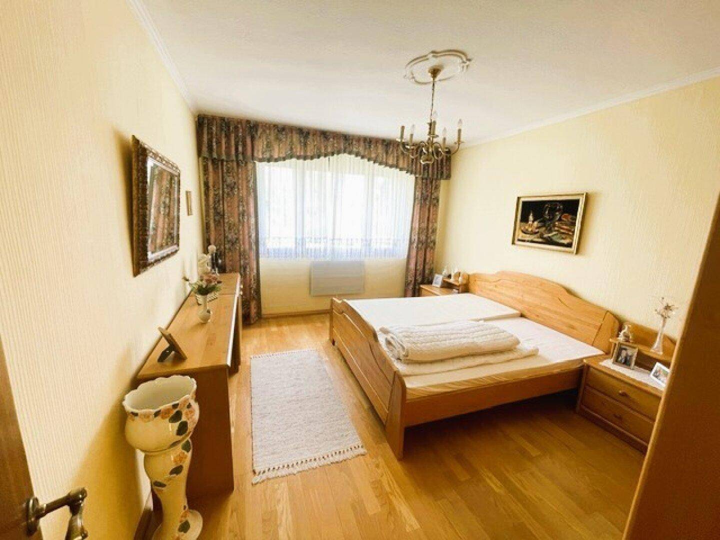 Helles Schlafzimmer mit einem Holzbett, Parkettboden und einer Stuckdecke mit Kronleuchter.