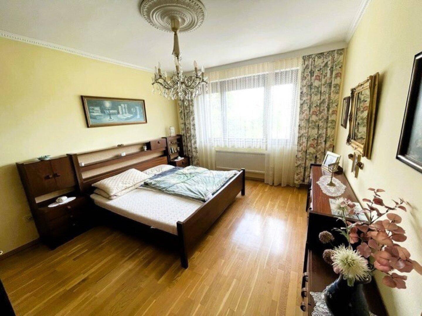 Schlafzimmer mit dunklem Holzbett, Parkettboden, Stuckdecke und einem eleganten Kronleuchter.