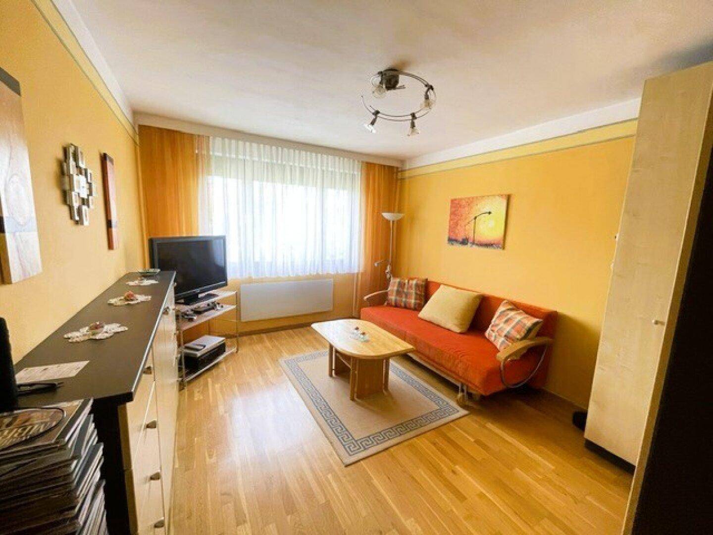 Gemütliches Wohnzimmer mit Parkettboden, einem orangefarbenen Sofa und großen Fenstern.