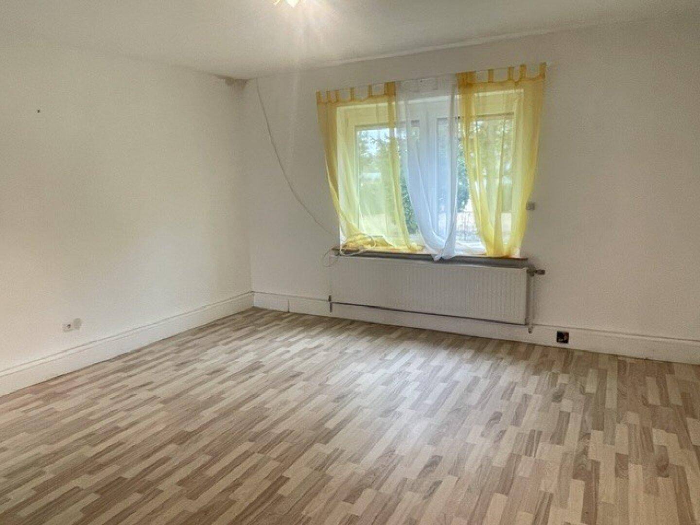 Leeres Zimmer mit Laminatboden, weißen Wänden und einem Fenster mit gelben Vorhängen.