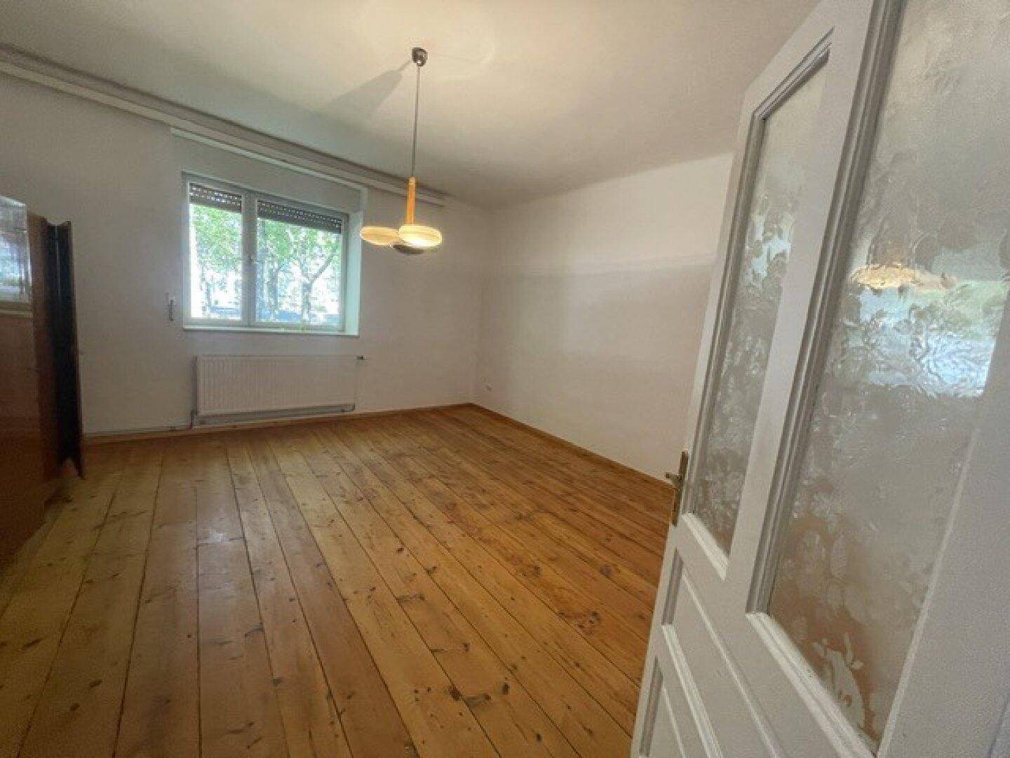 Geräumiger Raum mit Holzdielenboden, großem Fenster und einfacher Beleuchtung.