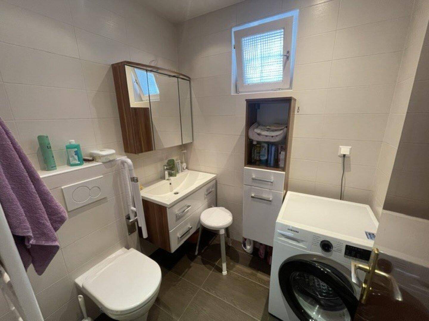 Modernisiertes Badezimmer mit weißen Fliesen, Waschmaschine und kleinem Fenster für Tageslicht.