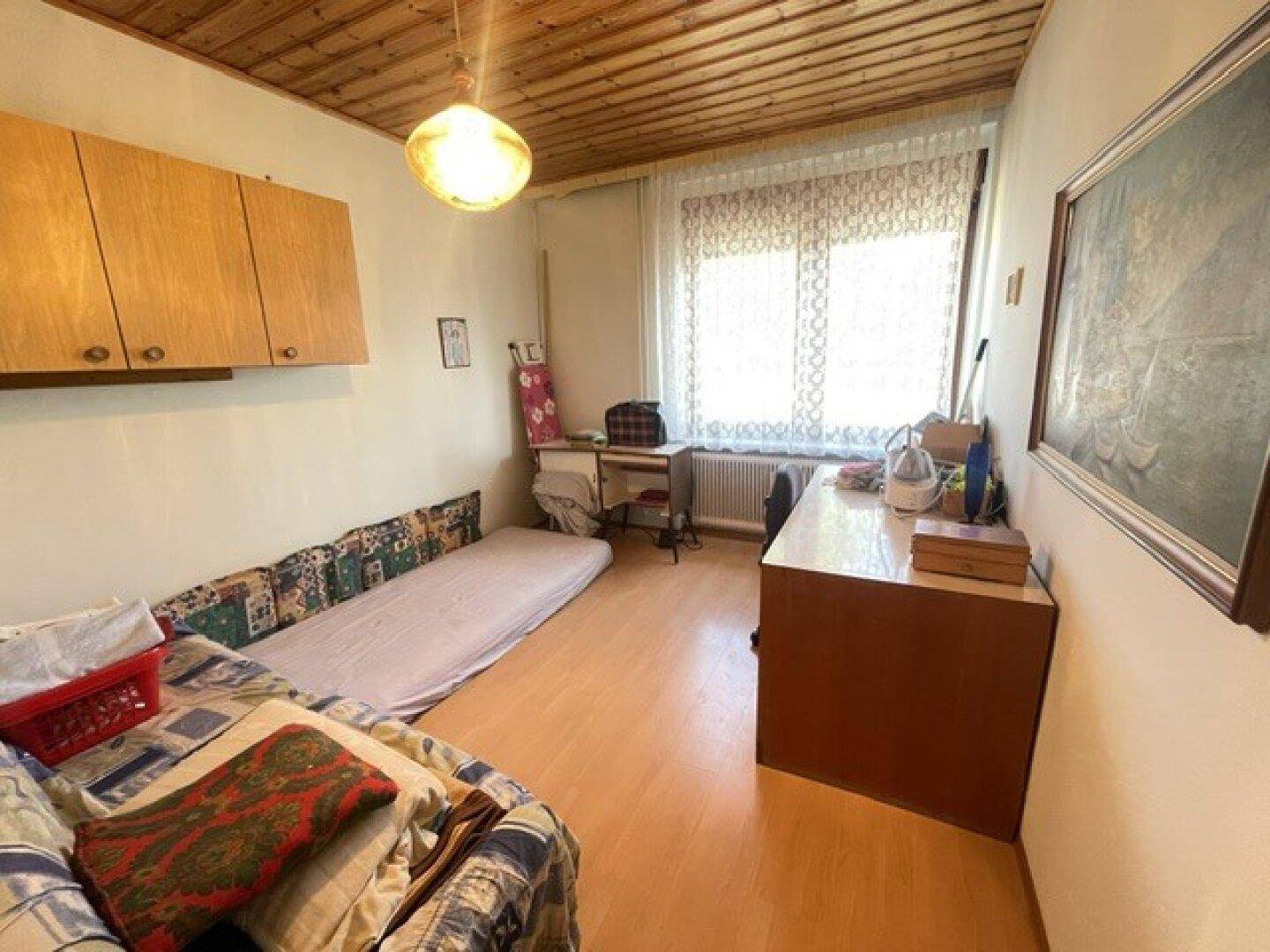 Einfaches Schlafzimmer mit Holzboden, Fenster und praktischen Holzschränken an der Wand.