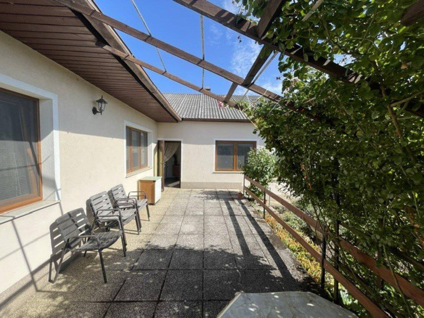 Große gepflasterte Terrasse mit Pergola und Sitzgelegenheiten, umgeben von üppigem Grün.