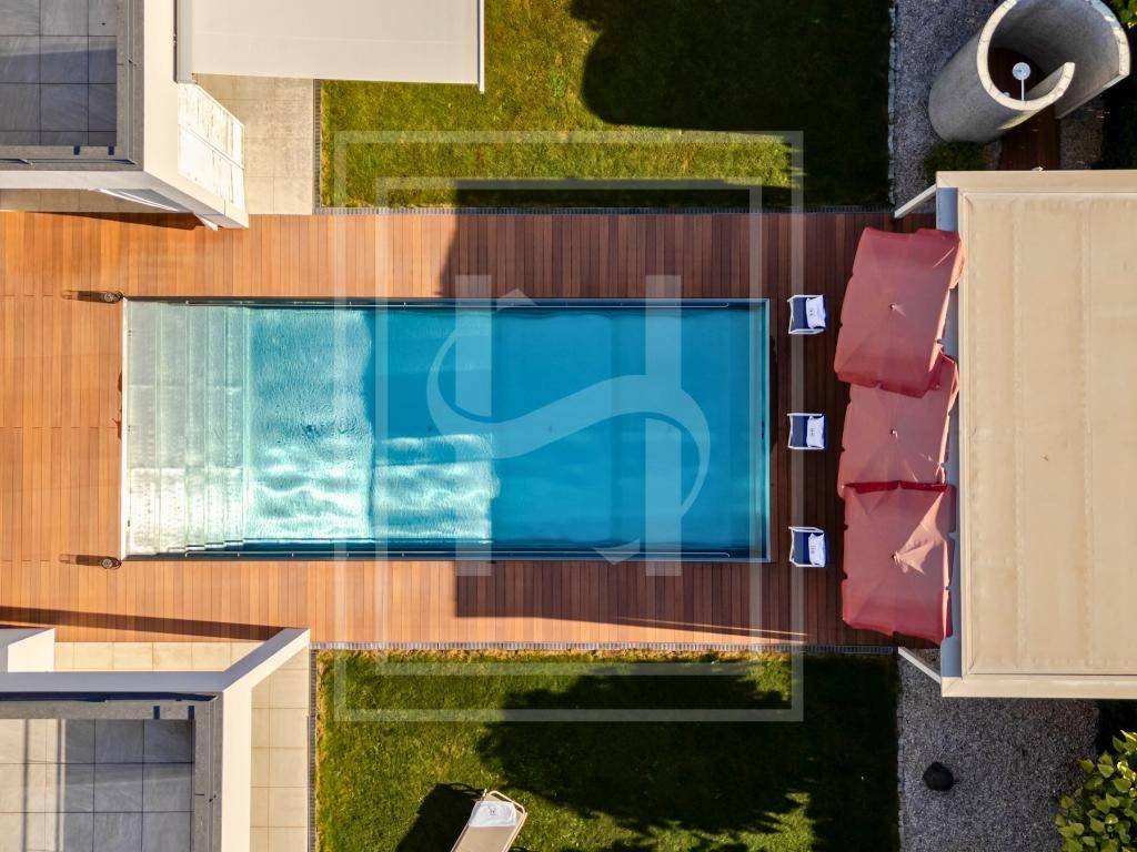 Vogelperspektive eines eleganten Pools mit Holzdeck, umgeben von gepflegtem Rasen und modernen Gebäudeteilen.