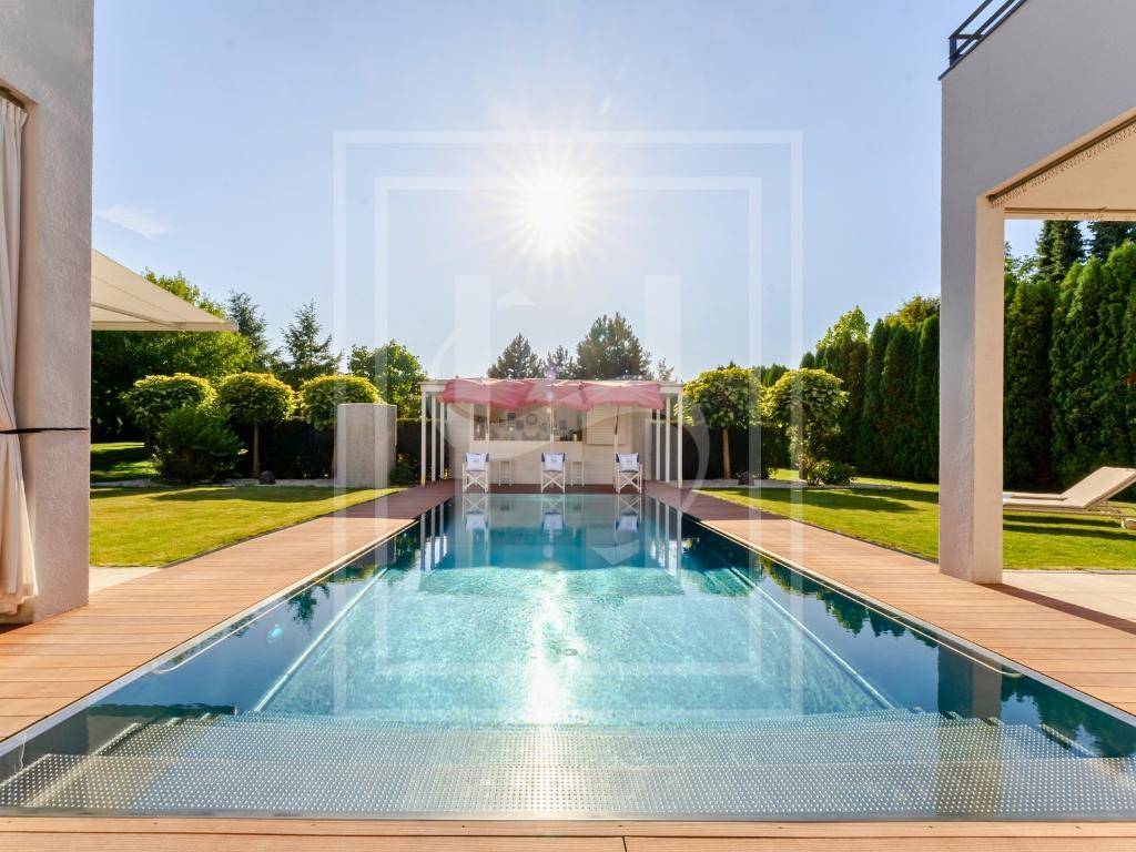 Langer Swimmingpool mit Holzdeck, umgeben von Rasen und einer stilvollen Poolbar mit roten Sonnenschirmen.
