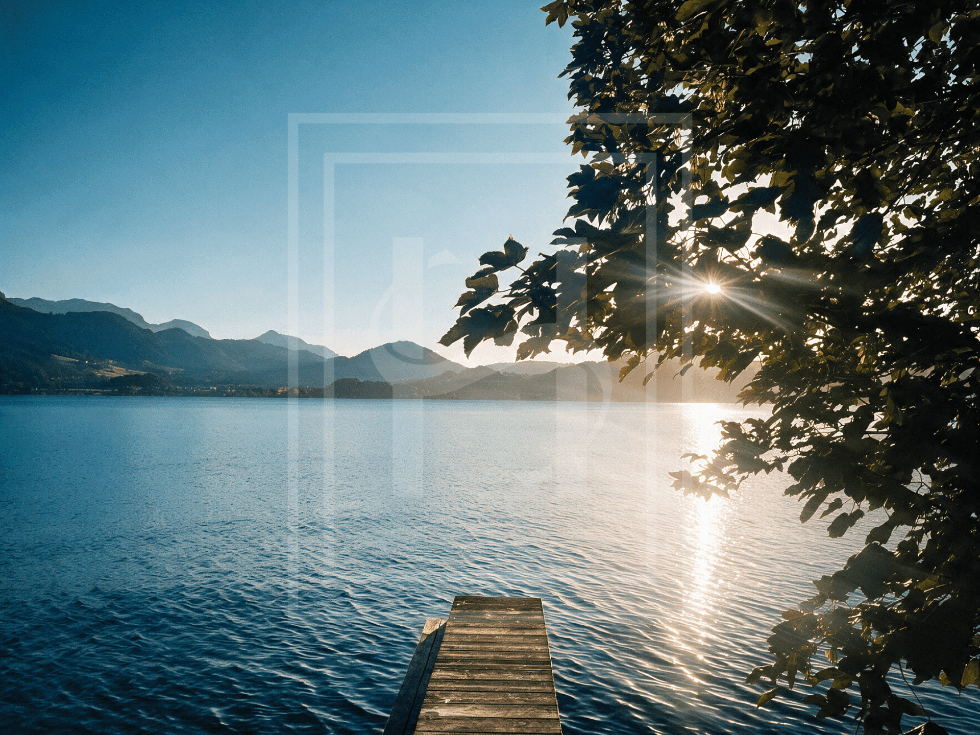 Sonnenuntergang über dem See mit Holzsteg und malerischer Bergkulisse im Hintergrund, durch Bäume gerahmt.