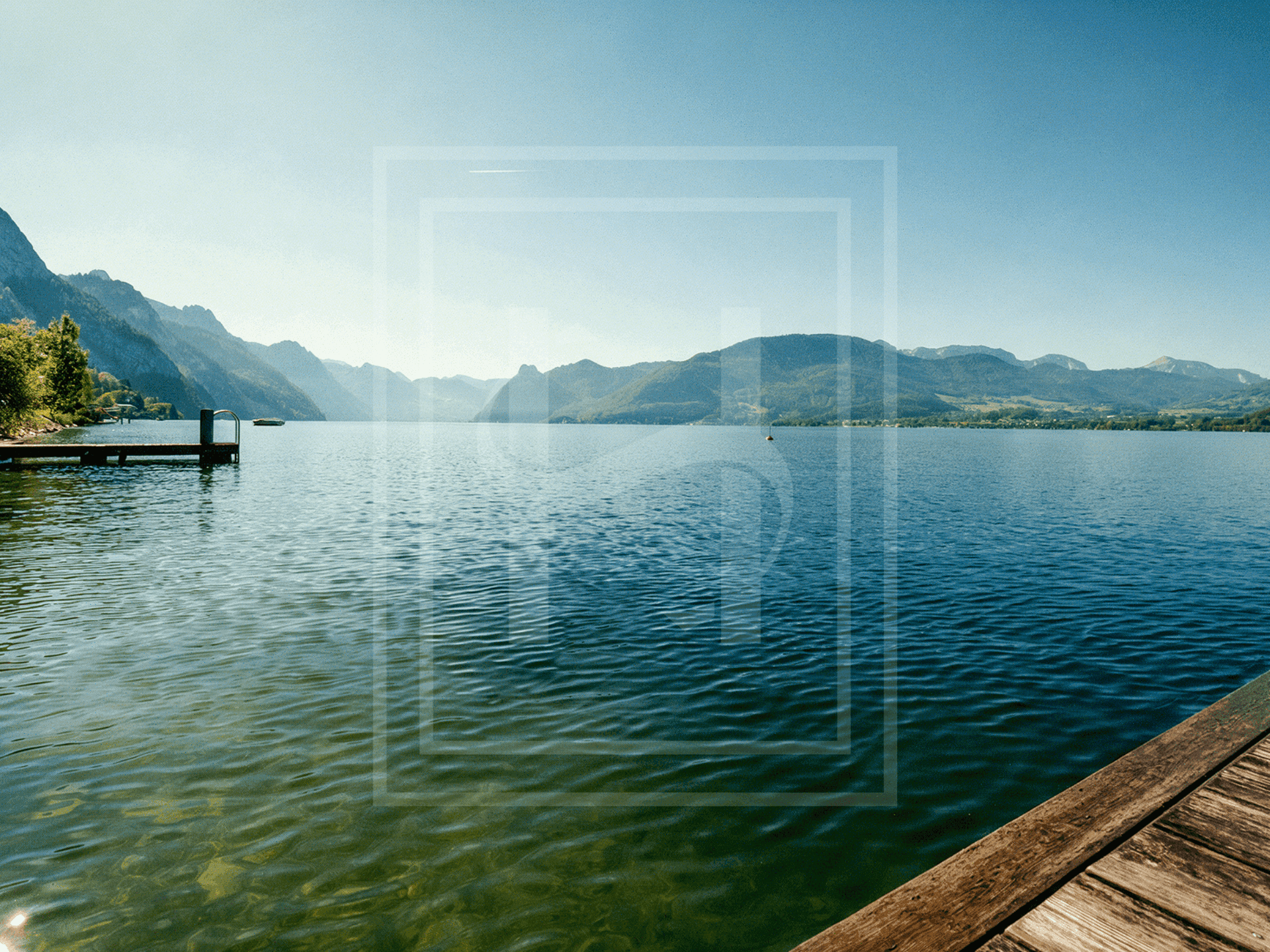 Klarer See mit weitem Blick auf die umliegende Berglandschaft und Holzstege unter strahlendem Himmel.