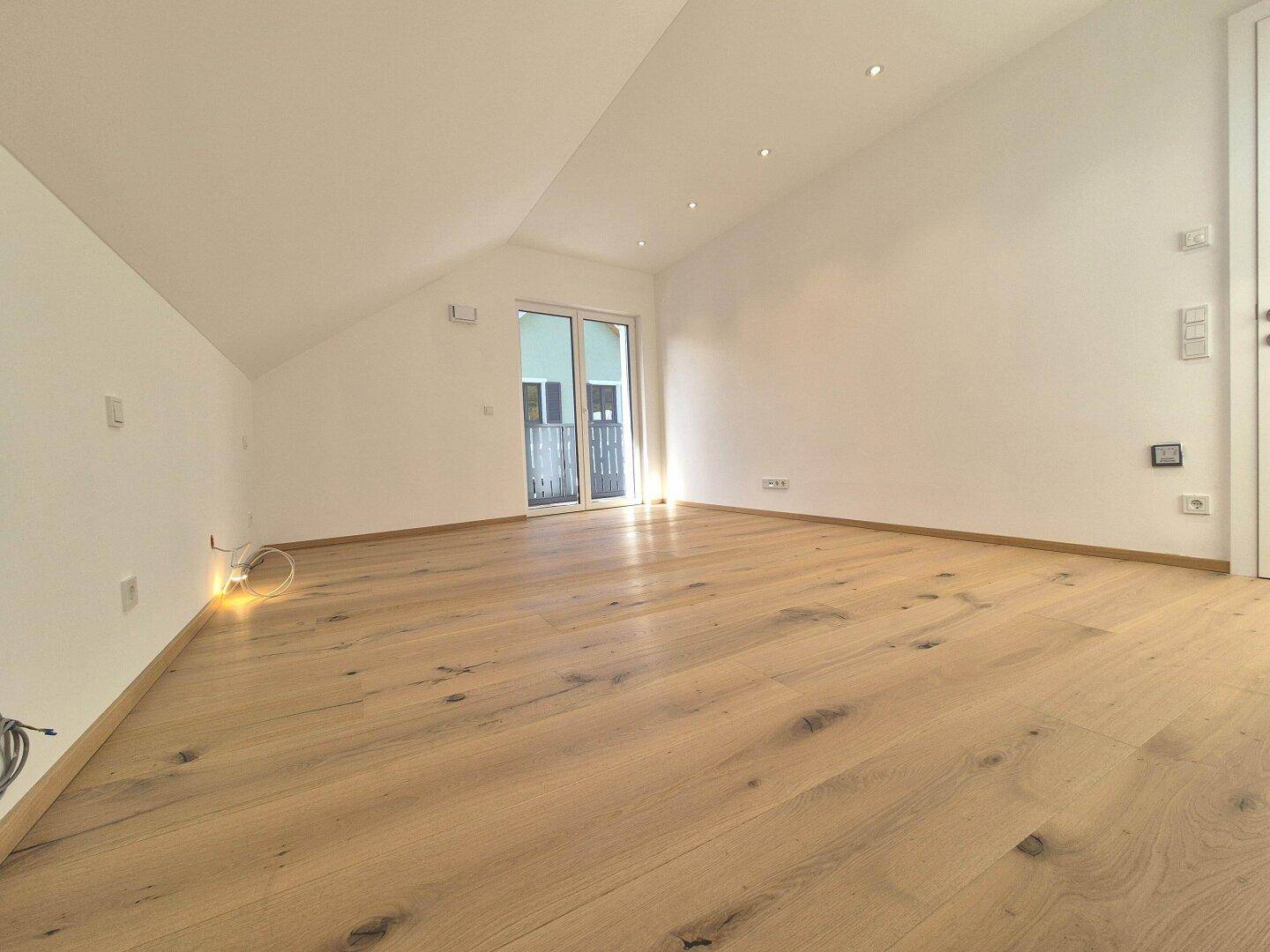 Geräumiges Zimmer mit hellem Holzparkett, Dachschrägen und viel Tageslicht durch die Balkontür.