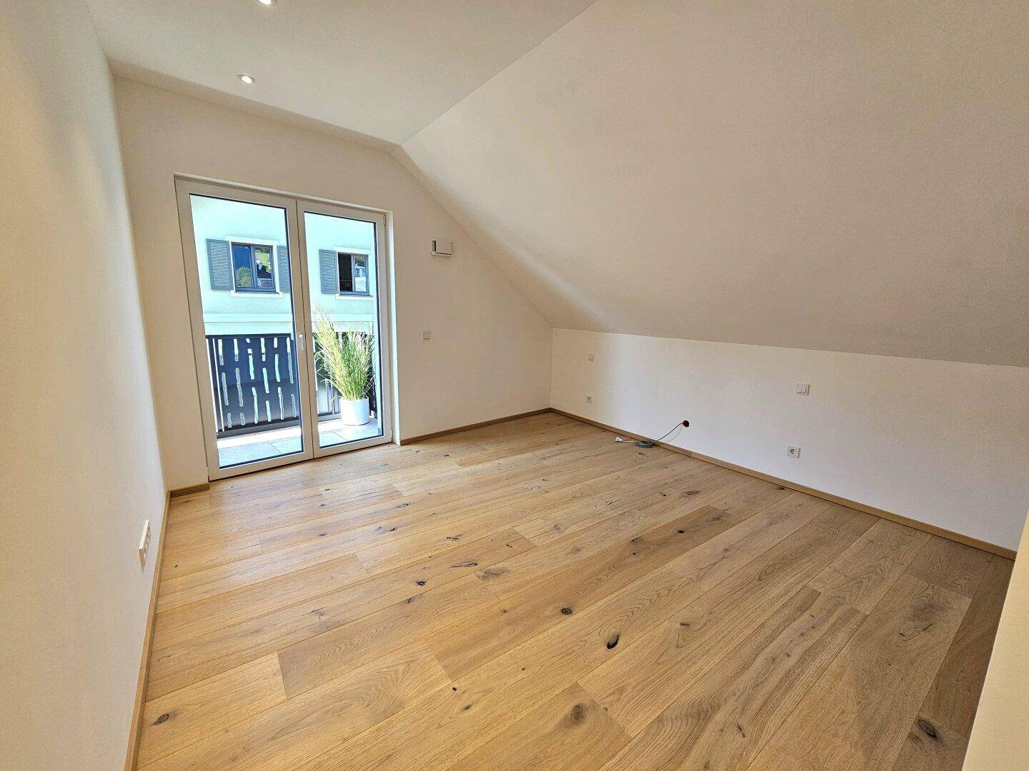 Geräumiges Zimmer im Dachgeschoss mit Holzboden und Zugang zu einem kleinen Balkon.