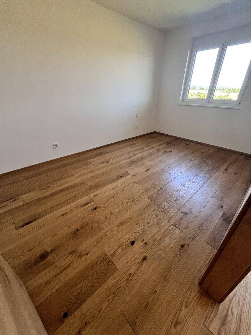 Helles, leeres Schlafzimmer mit Holzboden und großem Fenster für viel Tageslicht und Ausblick ins Grüne.