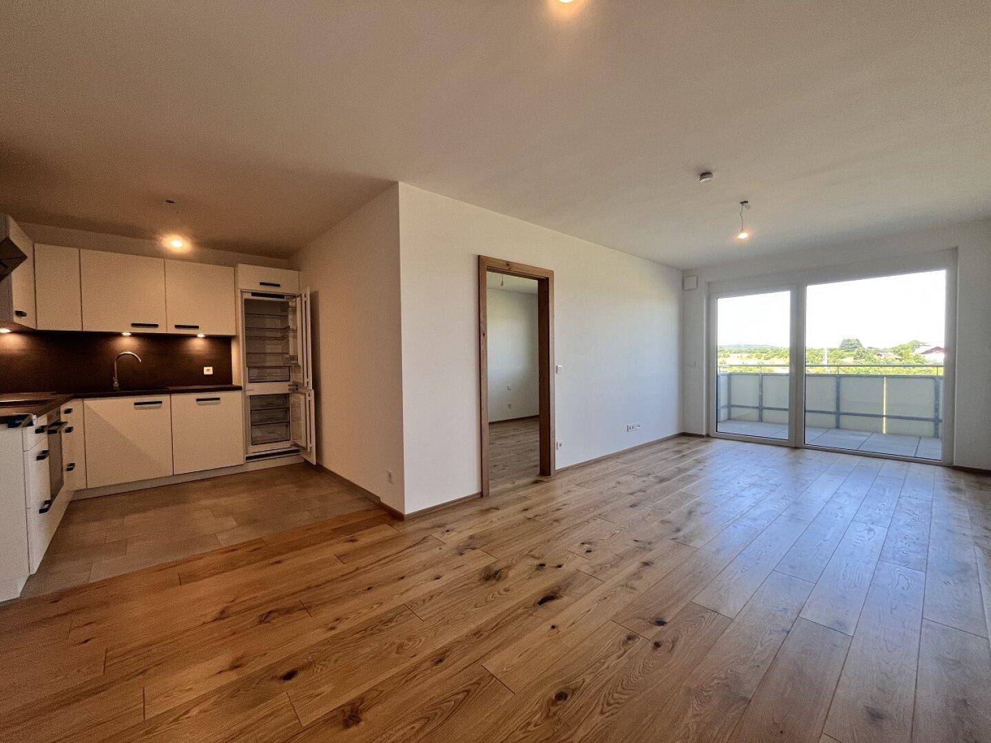 Geräumiger, offener Wohn-Essbereich mit moderner Küche und Zugang zum Balkon mit grüner Aussicht.