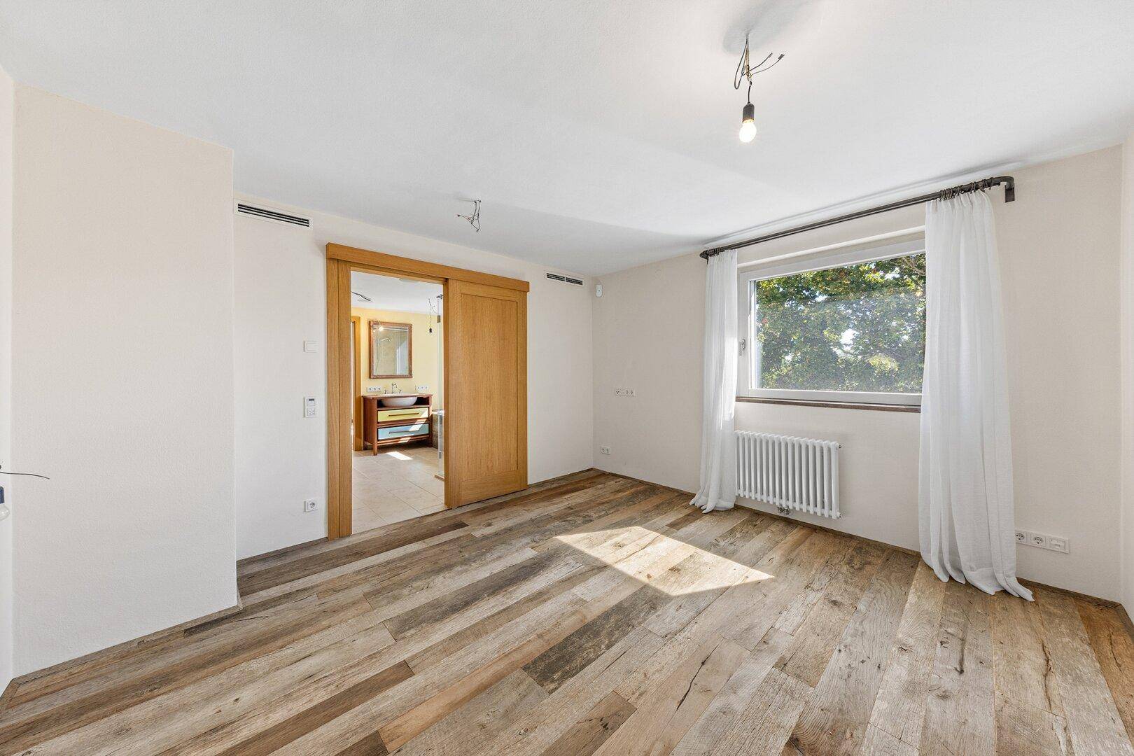 Helles Schlafzimmer mit rustikalem Holzboden und einem Fenster mit Blick ins Grüne.