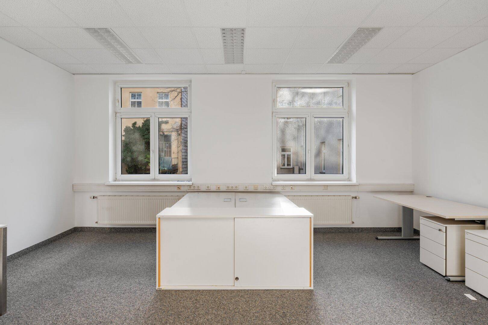 Geräumiges Büro mit zwei großen Fenstern, Teppichboden und schlichten Büromöbeln, bietet viel Tageslicht.