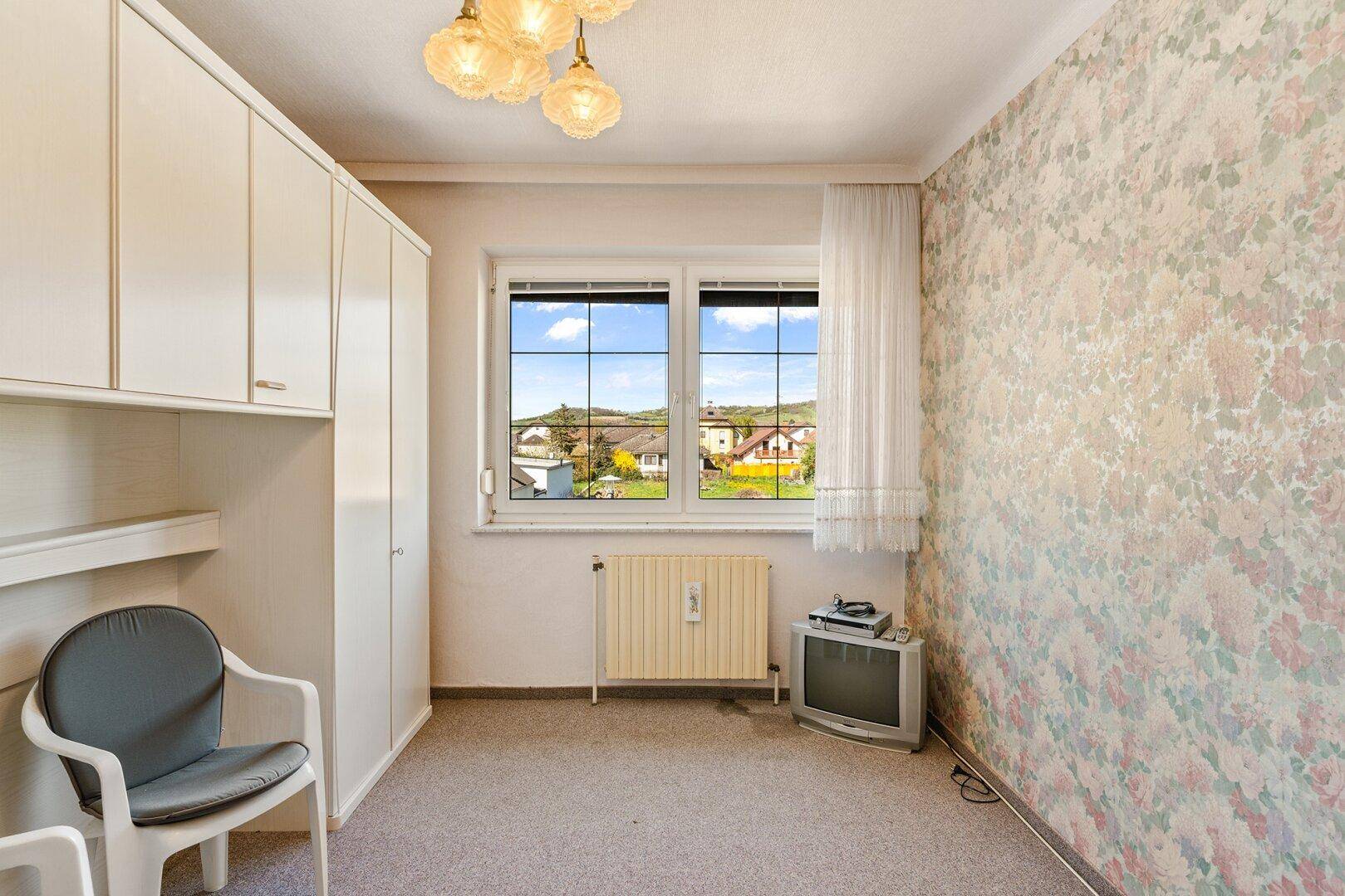 Zimmer mit floraler Tapete, Teppichboden und Fenster mit Blick ins Grüne.