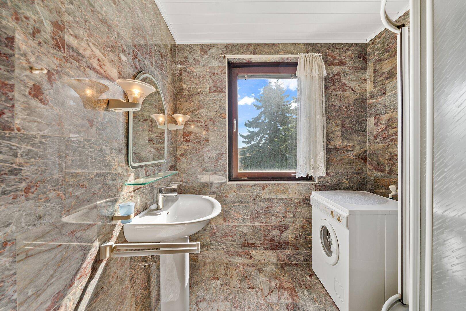 Badezimmer mit Dusche, Waschmaschine und Fenster, gefliest mit Natursteinoptik.