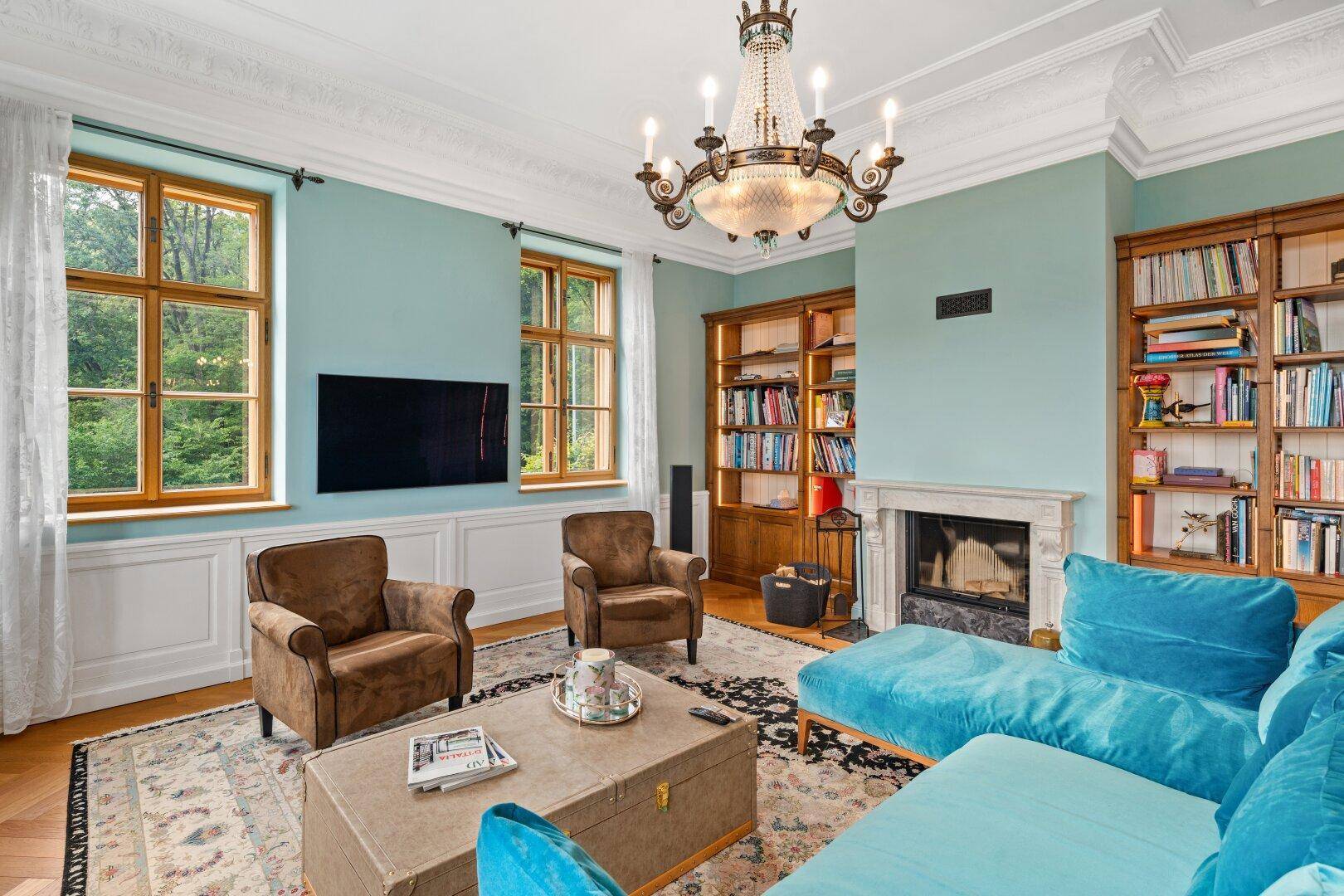 Gemütliches Wohnzimmer mit Kamin, blauer Couch und Blick ins Grüne durch große Fenster.