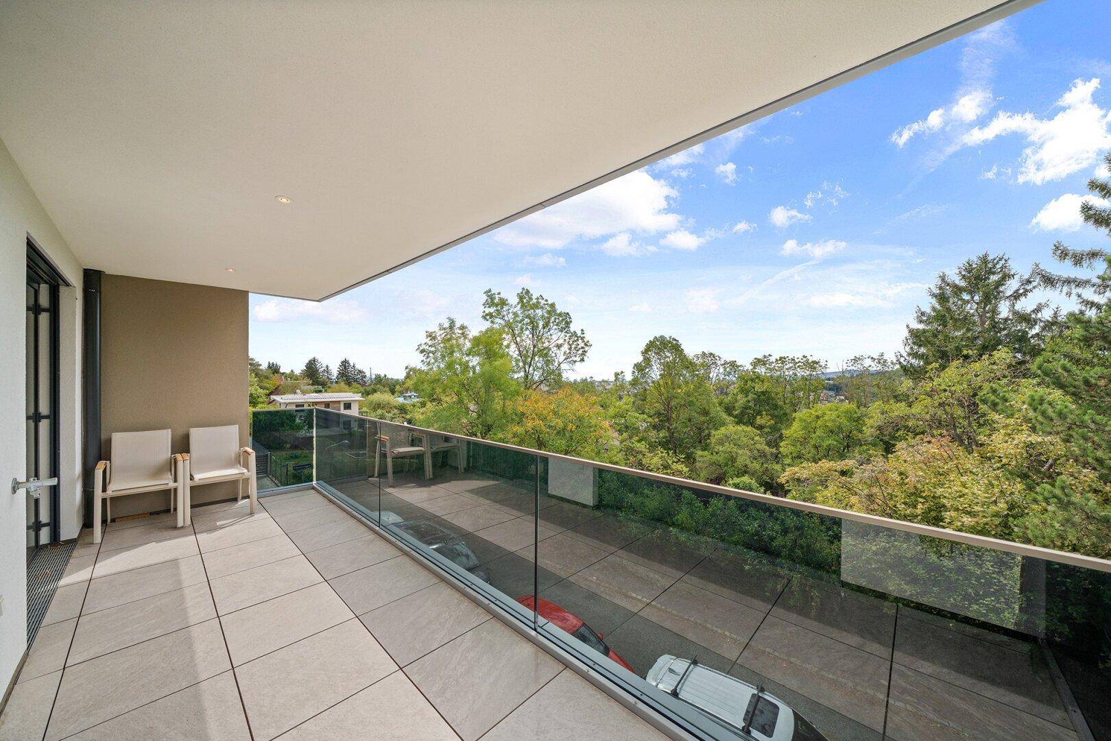Heller Balkon mit Glasgeländer, Sitzgelegenheiten und Panoramablick auf die umliegende Natur.