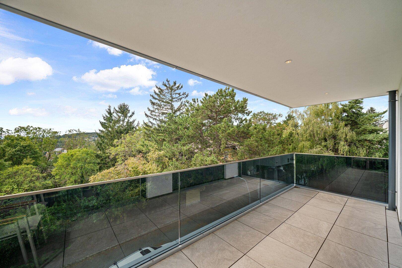 Geräumiger Balkon mit Glasgeländer, Fliesenboden und einem weiten Blick ins Grüne.