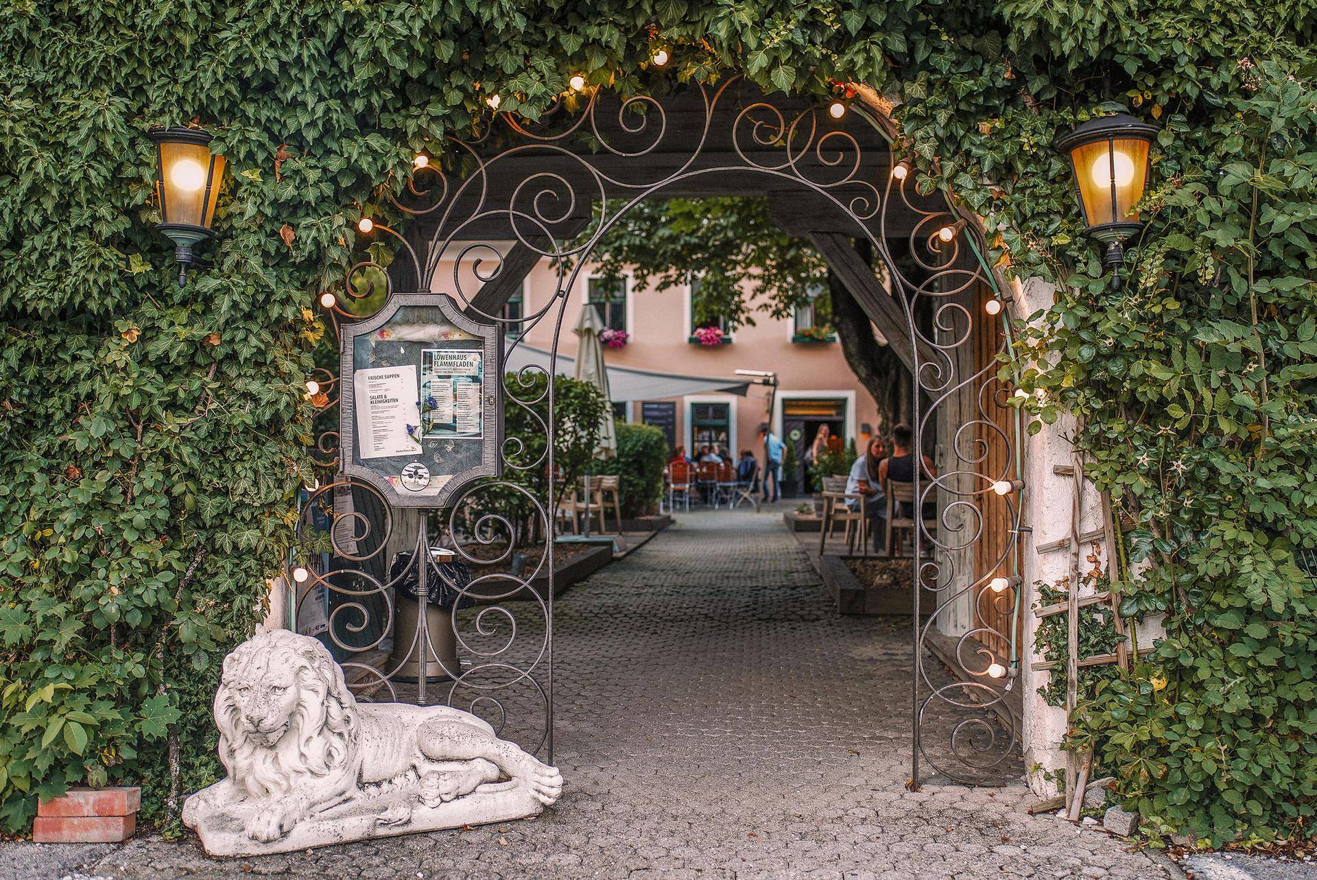 Einladender Eingang zu einem Innenhof mit Rankpflanzen, Lichterketten und einer steinernen Löwenstatue.
