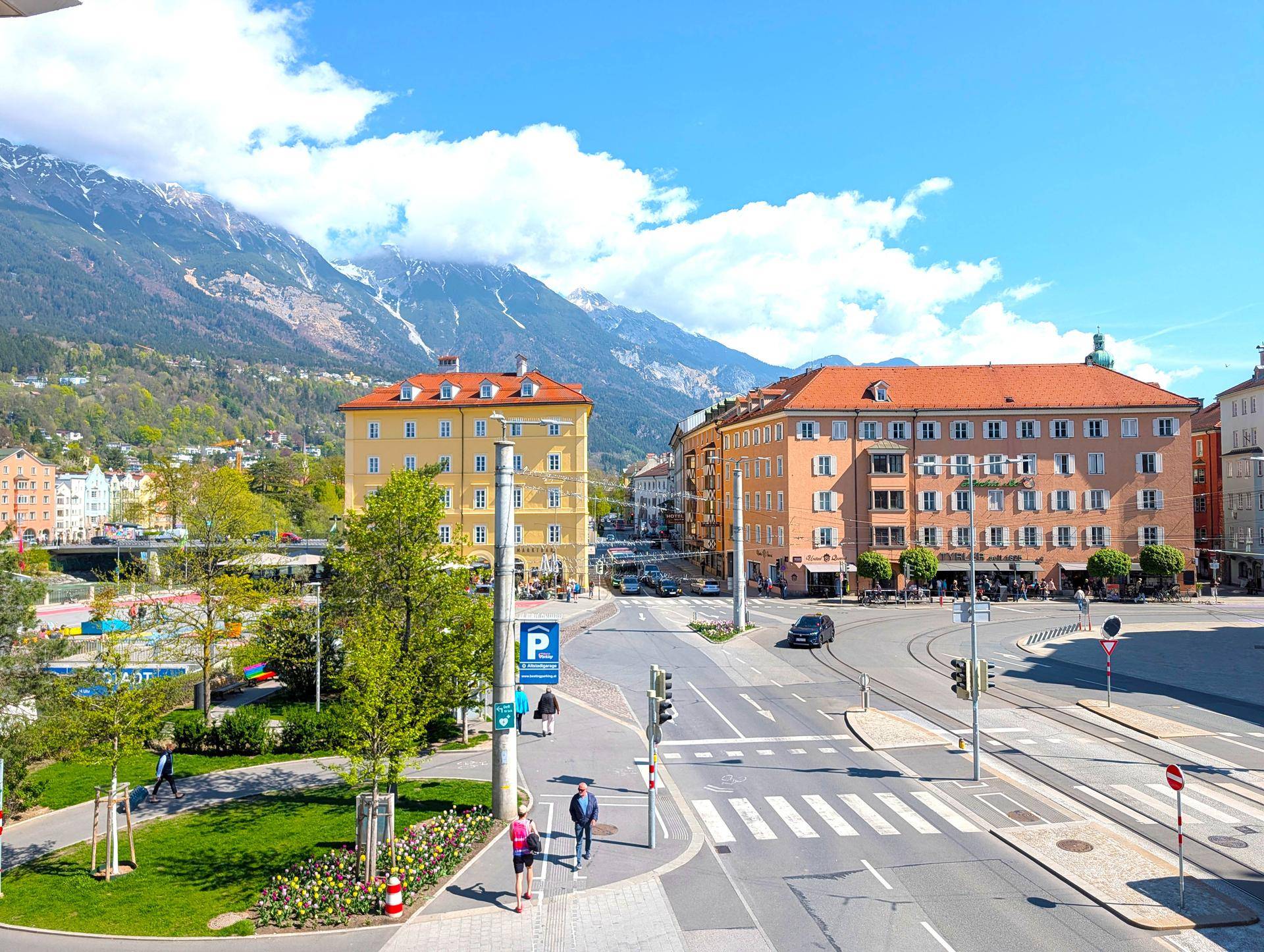 Panoramablick auf eine belebte Stadtstraße mit historischen Gebäuden und schneebedeckten Bergen im Hintergrund.