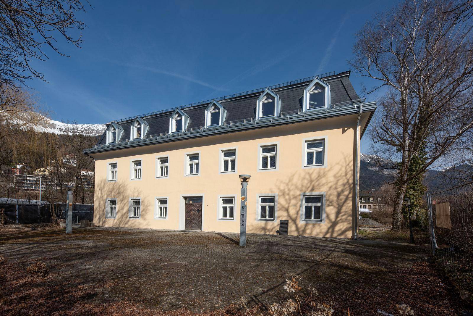 Vorderansicht eines sanierten Gebäudes mit gelber Fassade und Mansarddach, umgeben von Natur.