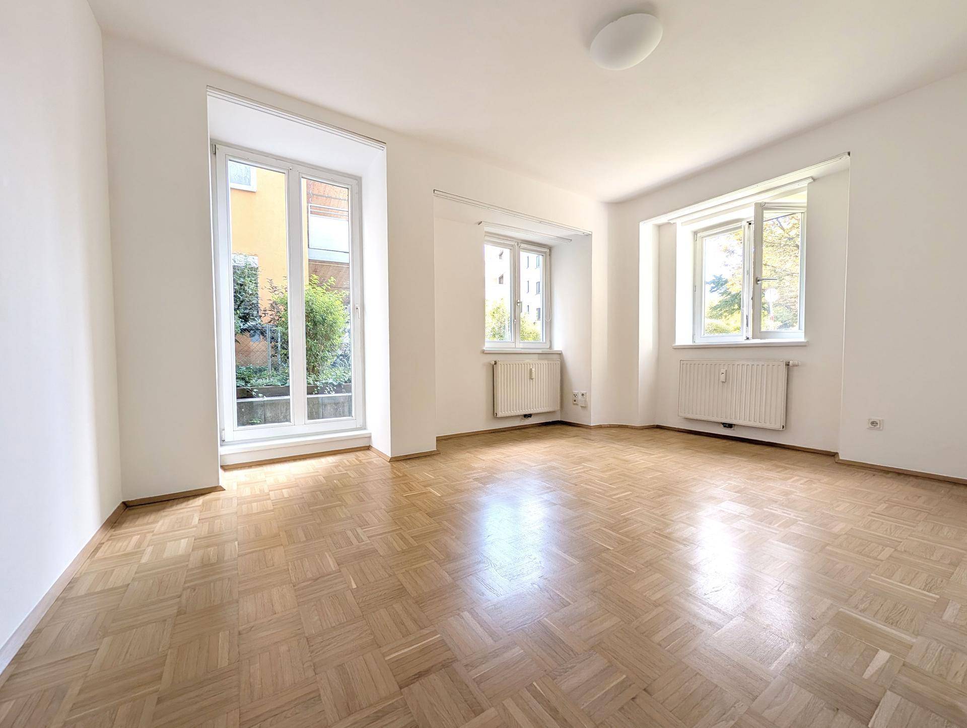 Helles, leeres Zimmer mit Parkettboden und großen Fenstern, die viel Tageslicht hereinlassen.