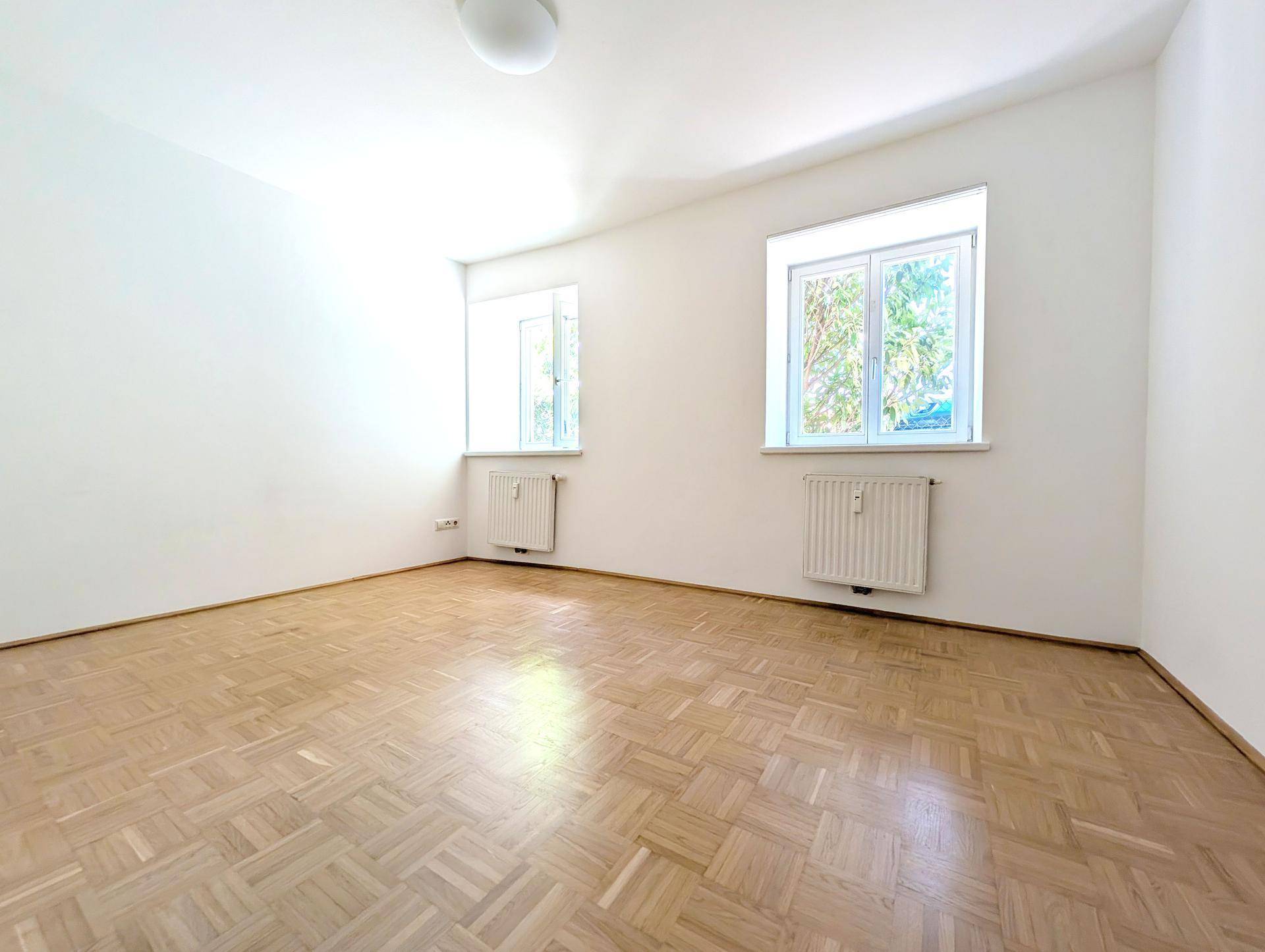 Leeres Schlafzimmer mit hellem Parkettboden und zwei Fenstern, die den Raum erhellen.