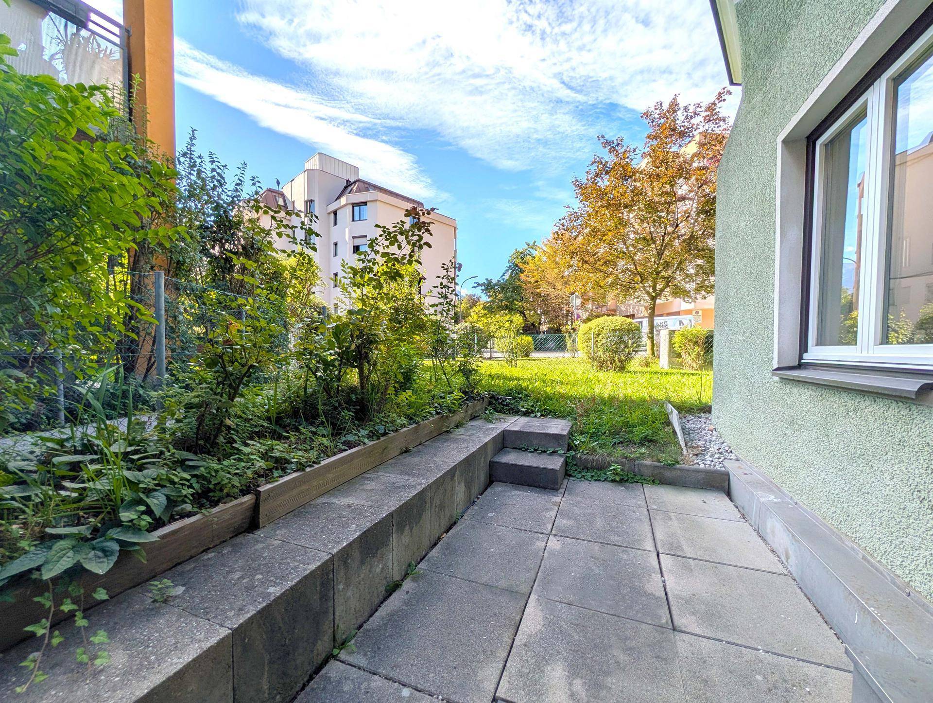 Gepflasterte Terrasse mit Stufen und Pflanzenbeeten, umgeben von Grünflächen und Gebäuden.