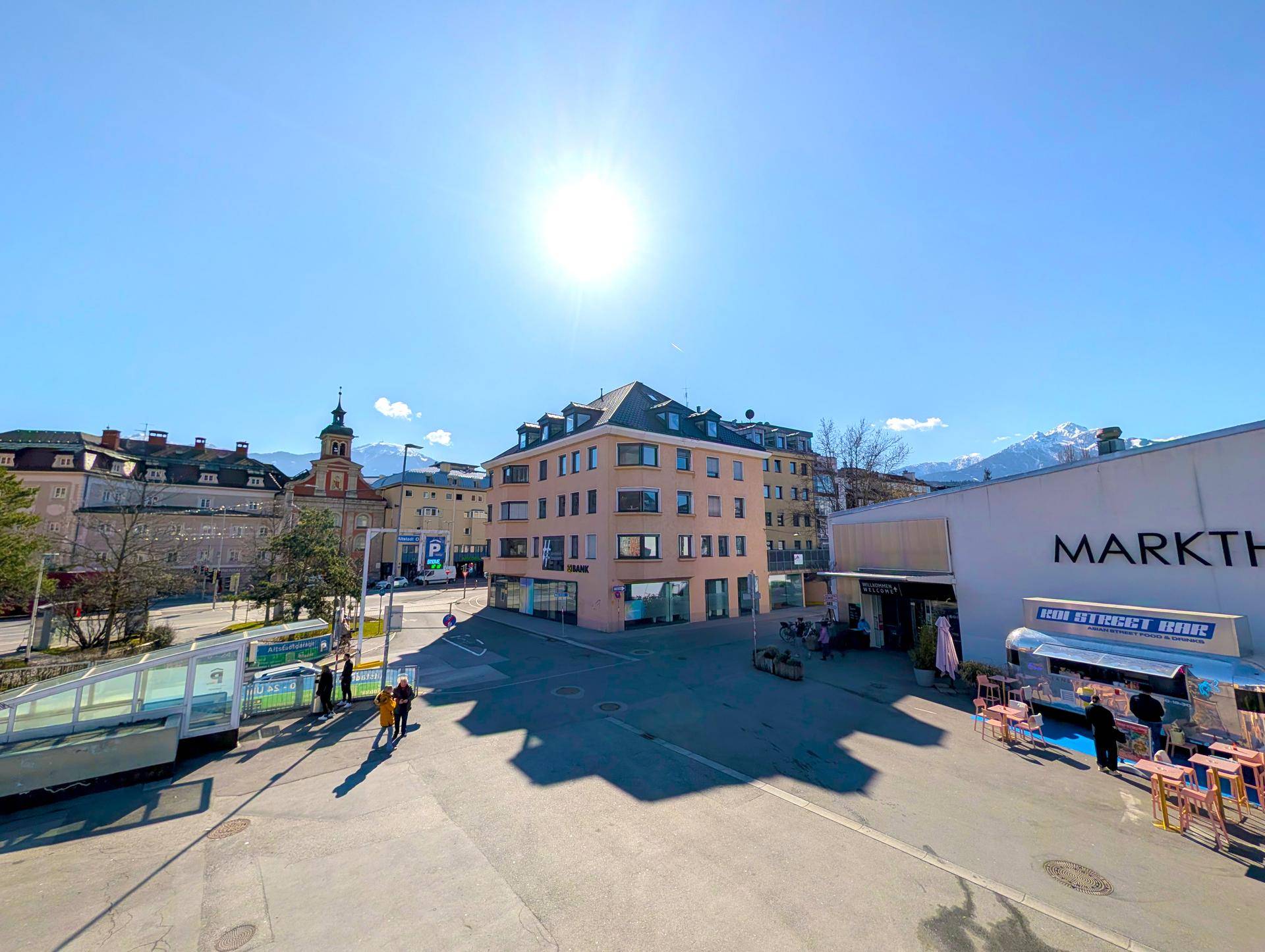 Sonniger Stadtplatz mit Gebäuden, einem Marktbereich und Bergen am Horizont.