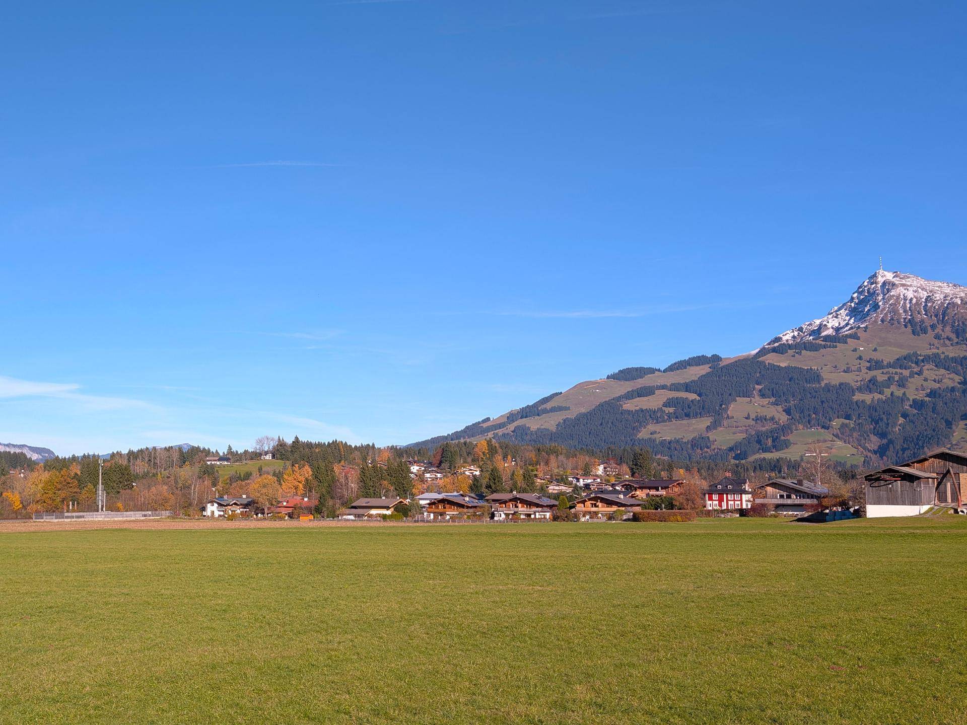 Weitläufige grüne Felder mit einem Dorf und schneebedeckten Bergen im Hintergrund unter blauem Himmel.