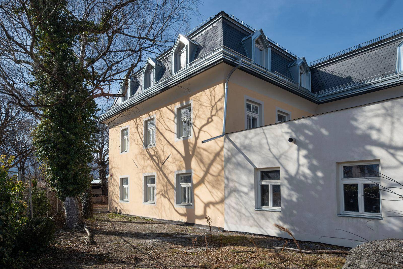 Seitenansicht eines Gebäudes mit gelber und weißer Fassade, umgeben von Bäumen und Schattenwurf.