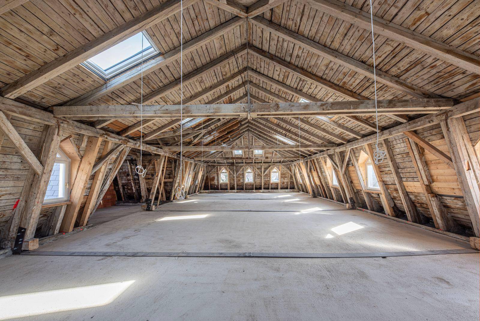 Großer, unfertiger Dachbodenraum mit sichtbaren Holzbalken, Dachfenstern und Betonboden.