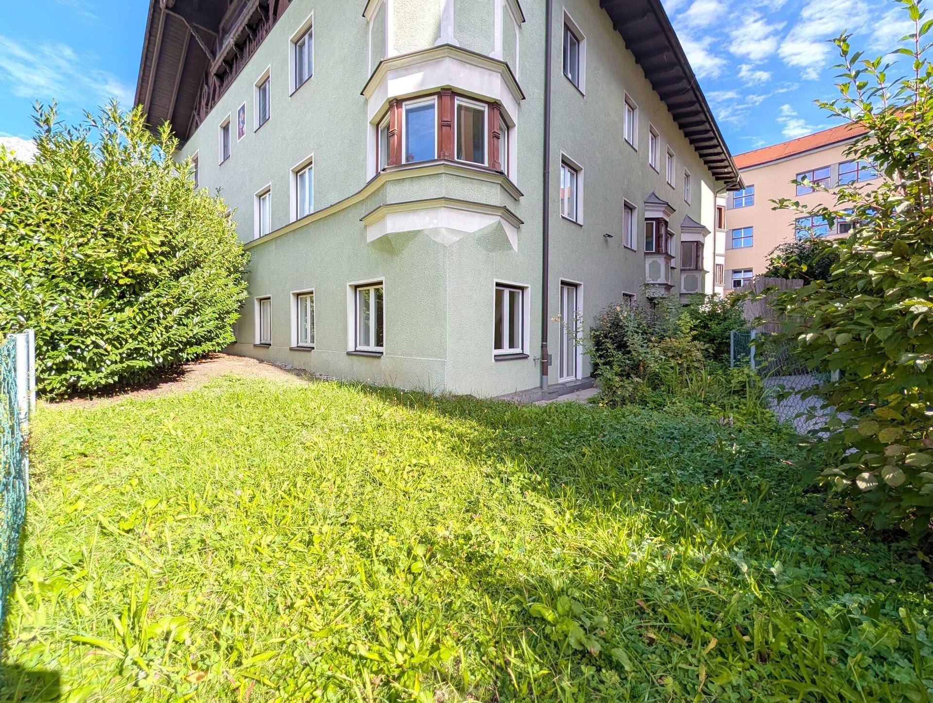 Grüner Gartenbereich neben dem Gebäude mit Rasenfläche und Büschen unter blauem Himmel.
