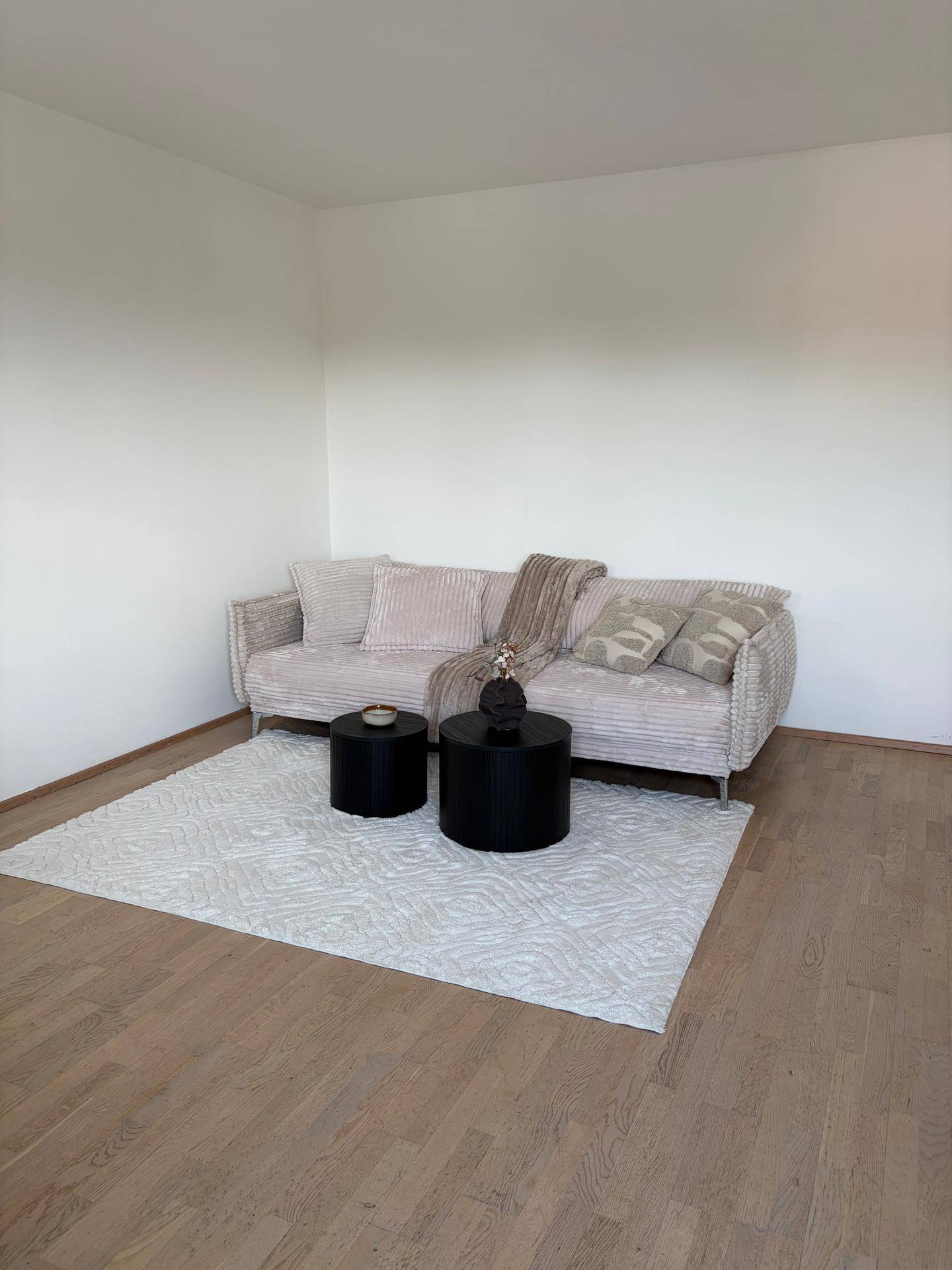 Geräumiges Wohnzimmer mit beigem Sofa, runden schwarzen Tischen und hellem Teppich auf Parkettboden.