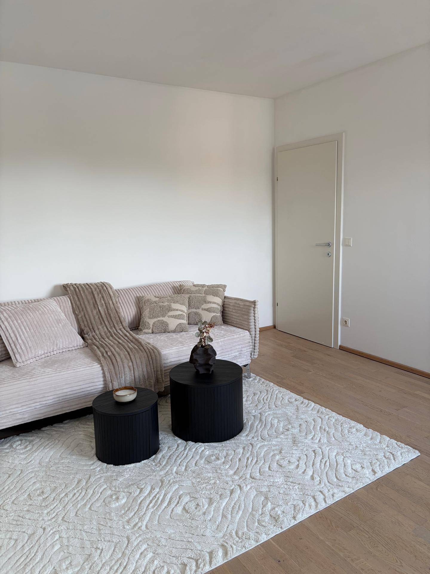 Gemütliches Wohnzimmer mit hellem Sofa, zwei schwarzen Couchtischen und einem strukturierten Teppich.