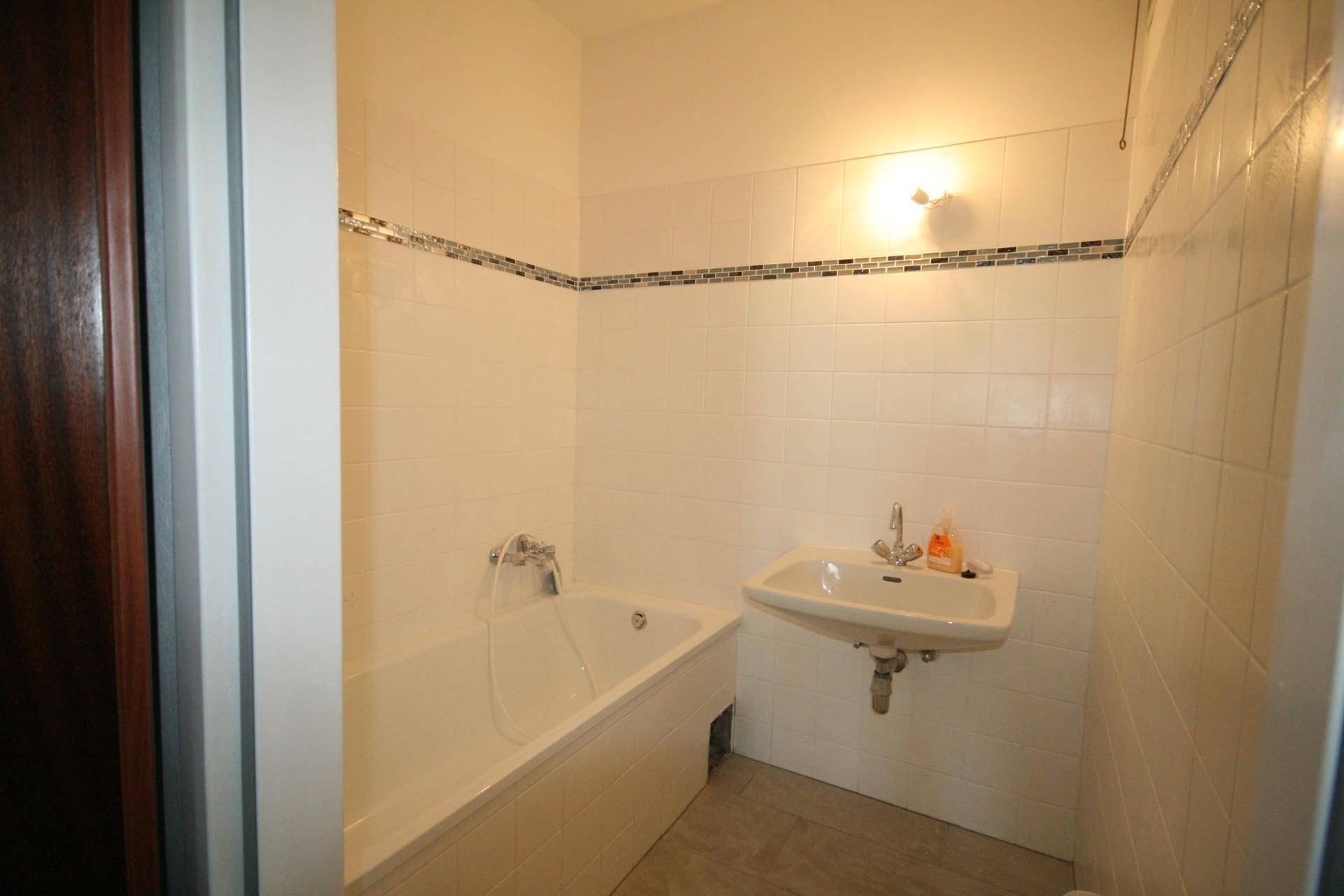 Funktionales Badezimmer mit Badewanne, Waschbecken und weißen Fliesen, akzentuiert durch eine Mosaikbordüre.