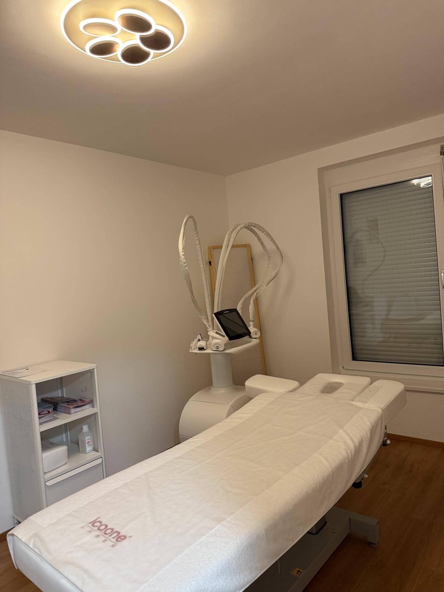 Heller Behandlungsraum mit Liege, moderner Schönheitsmaschine und Fenster mit Rollladen.