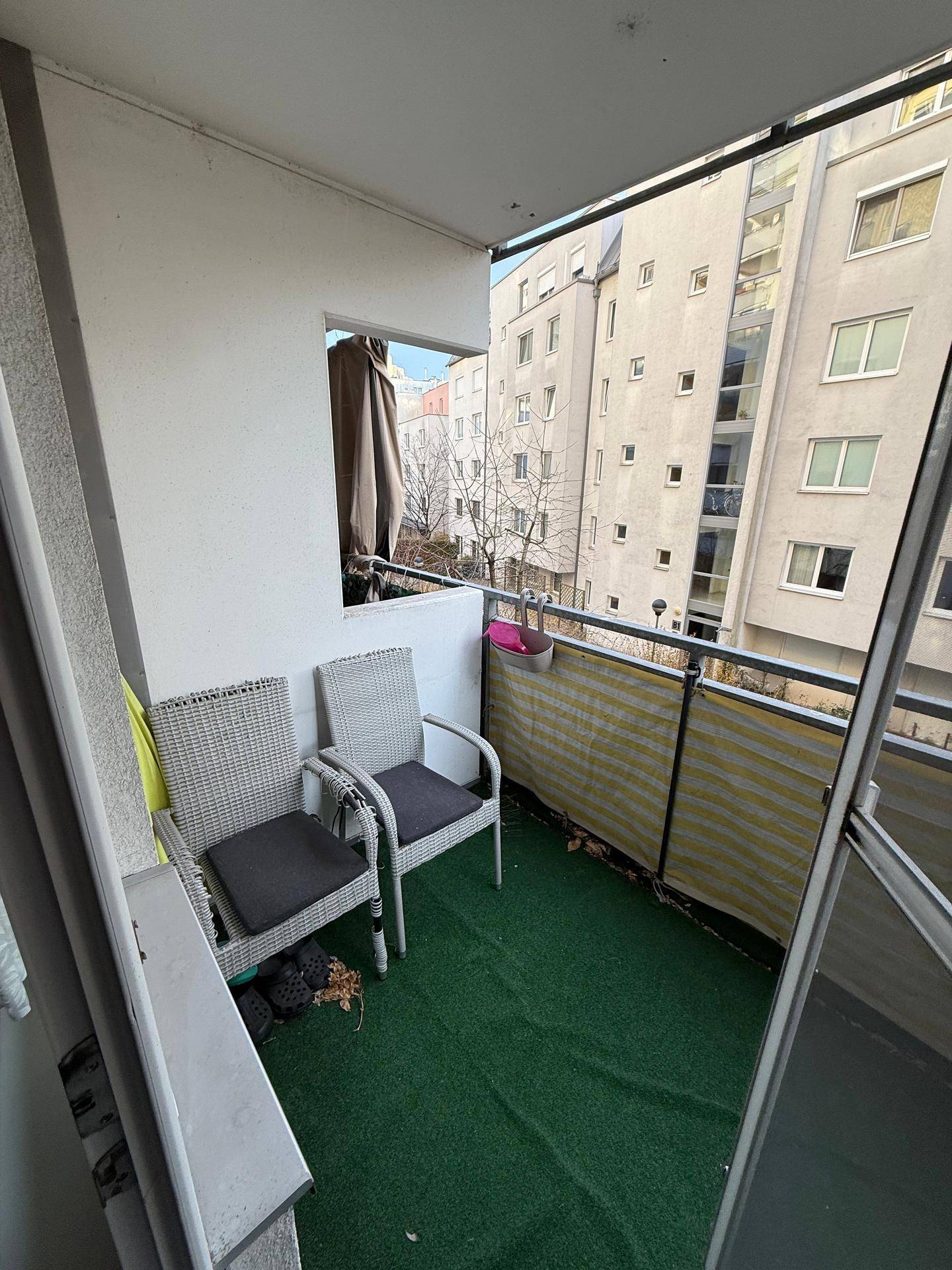 Gemütlicher Balkon mit Kunstrasen, Sitzgelegenheiten und Blick auf umliegende Gebäude.